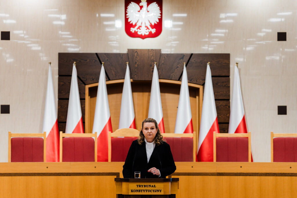 Kobieta przemawia przy mównicy w sali TK, na tle biało-czerwonych flag i godła