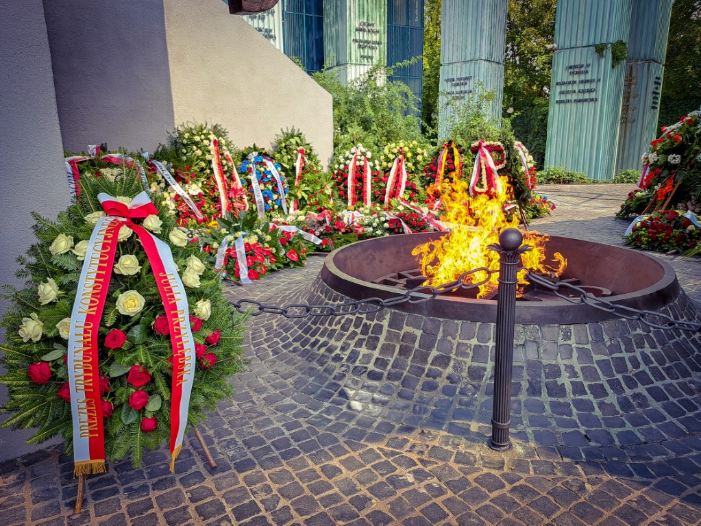 Płonący znicz wśród wieńców, z biało-czerwoną szarfą prezes TK po lewej stronie